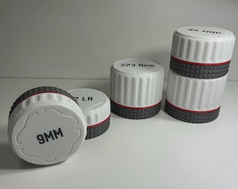 May include: A collection of four cylindrical white containers with ribbed sides and a textured dark gray base. Each lid features black printed text including 9MM, 22 LR, 223 Rem, and 44 MAG. A thin red ring separates the white top from the gray base.