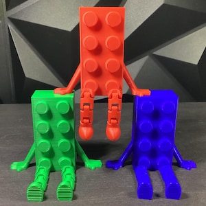 May include: Three 3D printed toy figures shaped like Lego bricks, sitting in a row. The figure on the left is green, the figure in the middle is red, and the figure on the right is blue.