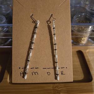 May include: A pair of long, beaded earrings with white and gold beads. The earrings are hanging from a brown card with the word "AMOR" written on it.