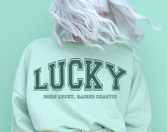 May include: A light green sweatshirt with the text "LUCKY" in a dark green arch above the text "BORN LUCKY, RAISED CHAOTIC" in a smaller font.