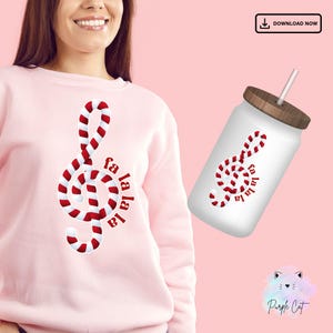Puede incluir: Una sudadera rosa con un diseño de bastón de caramelo rojo y blanco en forma de clave de sol. El texto "fa la la la" está escrito verticalmente junto a la clave.