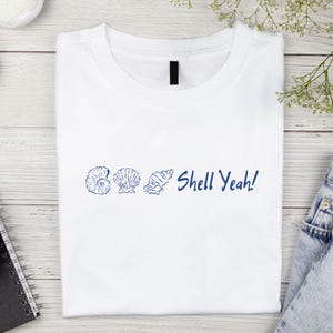 Puede incluir: Camiseta blanca con un diseño de conchas marinas azules y la frase "Shell Yeah!" impresa en la parte delantera. El diseño presenta tres ilustraciones diferentes de conchas marinas. La camiseta está hecha de un material suave y cómodo.