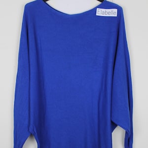 May include: A vibrant royal blue long-sleeve sweater featuring a wide boat neckline and a loose, draped batwing silhouette. The garment includes a small white rectangular tag near the neckline with the black printed text Elabelle.