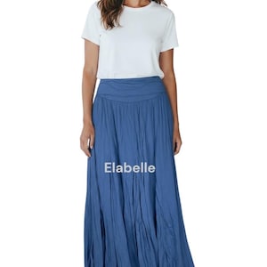 May include: A woman wearing a white t-shirt and a long, flowing blue skirt. The skirt has a gathered waistband and the text "Elabelle" is visible on the skirt. The outfit is paired with white sneakers.