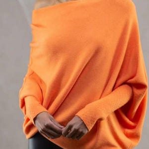 Puede incluir: Un suéter naranja brillante de hombros descubiertos con mangas largas. El suéter está hecho de una tela suave y de punto y tiene un ajuste holgado.