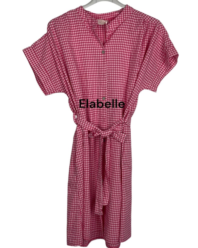 May include: A pink and white gingham dress with short sleeves and a matching belt. The dress has a button-down front and a mandarin collar. The text "Elabelle" is visible on the front of the dress.