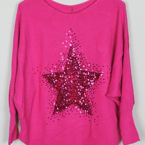 May include: A vibrant pink jumper featuring a sequined star design. The jumper has a round neck and long sleeves. The star is a prominent feature on the front of the garment.