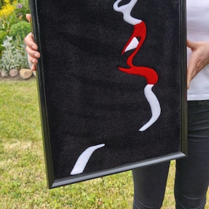 Puede incluir: Obra de arte enmarcada que presenta un perfil de rostro con labios rojos y detalles blancos sobre un fondo negro. La obra está en un marco negro y la sostiene una persona. La pieza se exhibe al aire libre sobre un césped.