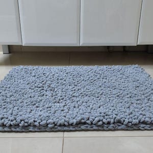 May include: A rectangular, light blue bath mat with a textured, looped surface. The mat is placed on a tiled floor in front of a white cabinet with silver legs. The mat's dimensions are not visible.