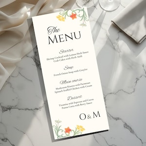 Puede incluir: Una tarjeta de menú color crema con detalles florales y escritura elegante. El menú enumera entrantes, sopas, platos principales y opciones de postre, incluyendo cóctel de camarones, sopa de cebolla francesa y risotto de champiñones. Sobre una superficie de mármol.