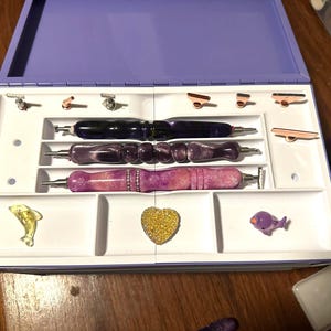 May include: A purple and white storage case with three purple and silver pens, a gold heart, a purple dolphin, and a few small silver and gold tools.