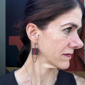 May include: Geometric leather earrings with a dark red and brown color scheme. The earrings feature a rectangular design with a chain detail. The jewelry is worn by a person.