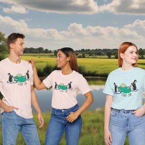 Könnte beinhalten: Drei Personen tragen weiße, rosafarbene und hellblaue T-Shirts mit einem Motiv von zwei Cartoon-Fröschen, die Zylinder tragen und auf einem Seerosenblatt sitzen. Der Text auf den Shirts lautet "C'mon Sir".