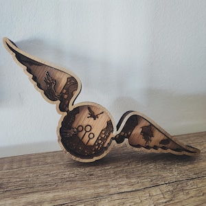 Può includere: Oggetto decorativo in legno raffigurante il Boccino d'Oro, con dettagli tagliati al laser. Il design presenta un centro rotondo con un paesaggio urbano e cerchi di Quidditch, affiancato da due ali. Un uccello vola al centro. In legno chiaro.