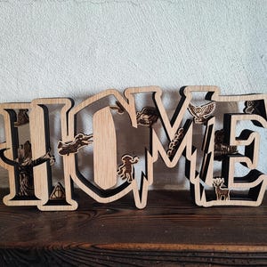 Op de afbeelding: Houten "HOME" bord met lasergesneden details. De letters zijn gevuld met iconische afbeeldingen, waaronder een hert, een uil en andere symbolen. Het bord is gemaakt van lichtgekleurd hout met een donkere rand en staat op een houten oppervlak.