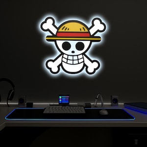 Könnte beinhalten: Leuchtende Wanddekoration mit dem Totenkopf- und gekreuzten Knochen-Emblem aus der Anime-Serie One Piece. Das Emblem ist weiß mit gelbem Hut und rotem Band. Darunter ein Schreibtisch mit Tastatur, Maus und Lautsprechern.