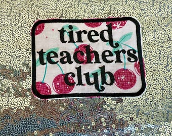 Op de afbeelding: Een rechthoekige patch met de woorden "tired teachers club" in zwart schrift. De patch heeft een witte achtergrond met rode kersenillustraties en groene bladeren, omlijnd in zwart. De achtergrond is een gouden paillettenstof.