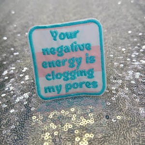 May include: Square patch with a white background, pink stripes, and a turquoise border. The patch reads "Your negative energy is clogging my pores" in turquoise lettering. The patch is on a sparkly silver surface.