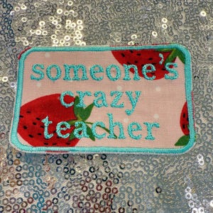 May include: A rectangular patch with a light pink background featuring red strawberries. The patch has a teal border and teal embroidered text that reads "Someone's crazy teacher".