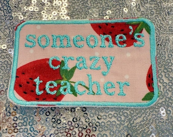 Op de afbeelding: Een rechthoekige patch met een lichtroze achtergrond met rode aardbeien. De patch heeft een turquoise rand en turquoise geborduurde tekst met de tekst "Someone's crazy teacher".