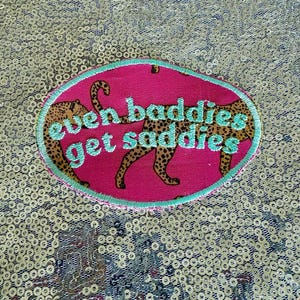 May include: Oval embroidered patch with the text "even baddies get saddies" in turquoise thread. The patch features two cheetahs with brown spots on a pink background, framed by a turquoise border.