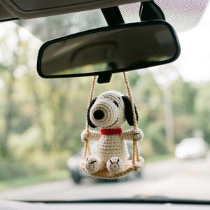 Könnte beinhalten: Ein gehäkelter Snoopy-Autoanhänger, der am Rückspiegel eines Autos hängt. Der Anhänger zeigt Snoopy auf einer Schaukel mit rotem Halsband und schwarzen Ohren. Der Hintergrund zeigt eine verschwommene Straße und Bäume.