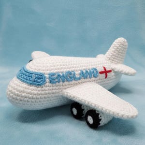 May include: A white crocheted airplane with blue accents and the word "ENGLAND" in blue. The airplane has a red cross on the tail and black wheels. The wings and tail are also white.