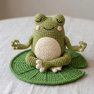 Könnte beinhalten: Gehäkelte Froschfigur in meditativer Pose, sitzend auf einem grünen Seerosenblatt. Der Frosch ist olivgrün mit geschlossenen Augen, rosa Wangen und einem cremefarbenen Bauch. Seine Arme sind in einem Yoga-Mudra ausgestreckt.
