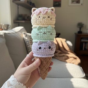 Könnte beinhalten: Eine handgefertigte gehäkelte Eistüte mit drei Eiskugeln in Form von Katzenköpfen. Die oberste Kugel ist hellbeige mit rosa Zuckerguss, die mittlere ist mintgrün und die untere ist lavendelfarben. Der Kegel ist braun.