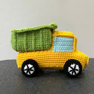 Op de afbeelding: Een gehaakte speelgoedkipper in geel met een groene laadbak en zwarte wielen met witte details. Het raam is lichtblauw. Een schattig, kleurrijk speelgoed.