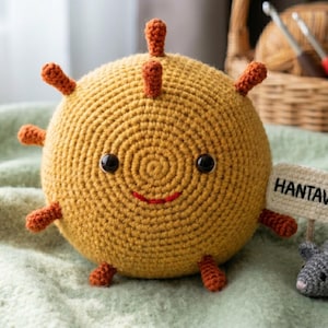 May include: A crocheted Hantavirus plushie with a smiling face, orange spikes, and a small gray mouse. The plushie is yellow and has a sign that says "Hantavirus". The background includes a basket with yarn and crochet hooks.