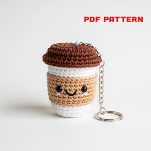 Puede incluir: Un llavero de taza de café de ganchillo con una cara sonriente. La taza es blanca, beige y marrón, con una tapa marrón. El llavero tiene una cadena y un anillo plateados. El texto en la imagen dice "PDF PATTERN".