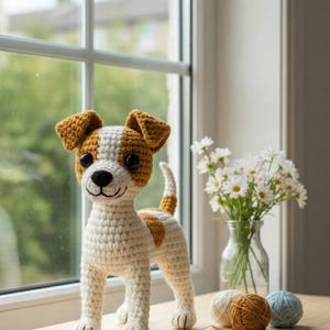 Op de afbeelding: Een handgemaakte gehaakte hondenfiguur in wit, bruin en beige, staand op een houten oppervlak. De hond heeft een lachend gezicht en een kleine staart. Een vaas met witte bloemen en bollen wol staan op de achtergrond.