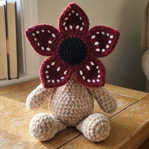 Puede incluir: Un peluche Demogorgon hecho a mano con ganchillo. El peluche presenta una cabeza en forma de flor de color burdeos con detalles blancos, un centro negro y un cuerpo, brazos y piernas beige. Está sentado sobre una mesa de madera.