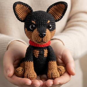 Puede incluir: Figura de perro de ganchillo con cuerpo negro, detalles marrones y collar rojo. El juguete hecho a mano se sostiene en manos abiertas, mostrando su detallada artesanía. Las orejas y patas del perro son marrones.