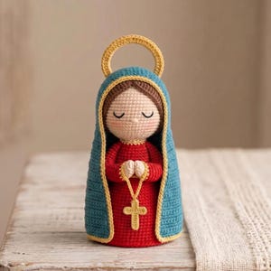 Op de afbeelding: Een gehaakte pop die een figuur met gesloten ogen voorstelt, gekleed in een rode jurk en een blauwe kapmantel met een gele rand. Het figuur heeft een gele halo boven het hoofd en houdt een gele gehaakte rozenkrans met een kruis vast.