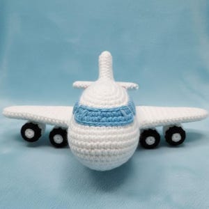 Könnte beinhalten: Ein handgefertigtes Häkel-Flugzeug in Weiß mit blauen Akzenten. Das Spielzeugflugzeug hat Flügel, ein Heck und vier schwarze Räder. Das Flugzeug steht auf einer hellblauen Oberfläche.