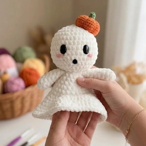 May include: A handmade white crochet ghost doll with black eyes, rosy cheeks, and a small orange pumpkin hat. The ghost has small arms and a flowing body. The doll is a cute, whimsical Halloween decoration.