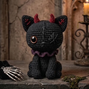 May include: A black crocheted cat plush toy featuring two small red horns, a purple ruffled collar, one large glossy brown eye, and one eye stitched with black thread. The ears have red cross-stitch detailing, and the body has visible seams.