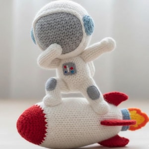 May include: A knitted astronaut doll in a white spacesuit with gray accents, standing on a red, white, and blue rocket. The astronaut has a gray helmet and blue ear pieces. The rocket has red fins and orange flames.