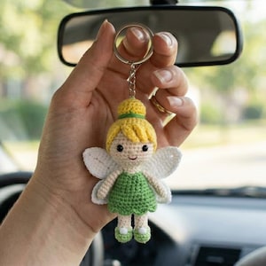May include: A crochet keychain featuring a small fairy figure with yellow hair in a bun, a green dress, and white wings. The figure has a light beige face with black bead eyes and a small stitched mouth, attached to a silver metal split ring.