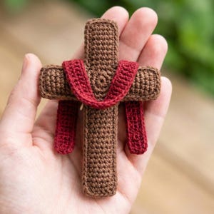 Könnte beinhalten: Ein handgefertigtes Häkelkreuz in Braun- und Rottönen. Das Kreuz wird in einer Hand gehalten, wodurch die detaillierten Nähte zur Geltung kommen. Das rote Garn ist über das Kreuz drapiert und fügt ein dekoratives Element hinzu.