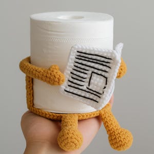 Puede incluir: Un rollo de papel higiénico blanco está envuelto en una funda de ganchillo de color amarillo mostaza, con brazos y piernas. El personaje de ganchillo sostiene un pequeño cuadrado blanco con líneas negras, que se asemeja a un periódico. El texto "PDF PATTERN" está en la parte superior.