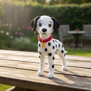 Puede incluir: Figura de perro dálmata hecha a mano con ganchillo. El perro blanco tiene manchas negras, orejas negras y un collar rojo con una etiqueta dorada. El perro está sobre una superficie de madera, con un fondo verde borroso.