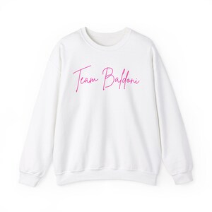 May include: White sweatshirt with a pink graphic that says "Team Baldoni".