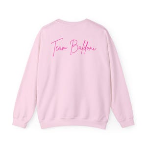 May include: Pink sweatshirt with the text "Team Baldoni" in pink lettering on the back.