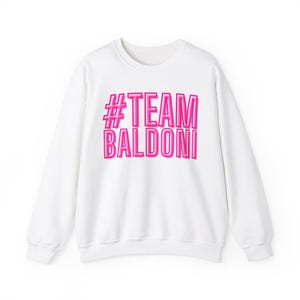 May include: White sweatshirt with a pink graphic that says "#TEAM BALDONI"