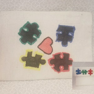 May include: A white canvas with four puzzle pieces painted in green, blue, brown, and brown. The puzzle pieces are arranged around a pink heart. The canvas is surrounded by a white border.