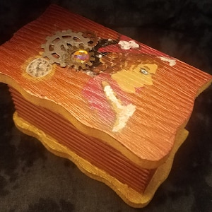 May include: A wooden box with a decorative design featuring a woman's face, gears, and a jewel. The box is painted in shades of brown, red, and gold.
