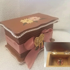 May include: A small wooden box with a brown painted finish and a pink ribbon. The box is decorated with painted flowers and a bee with gold glitter. The lid is open to reveal a small bee inside.